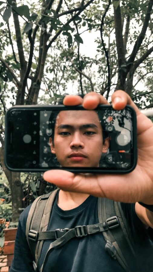 jovem olhando para a câmera através da tela do celular enquanto está na natureza, simbolizando equilíbrio e melhorar relação com a tecnologia