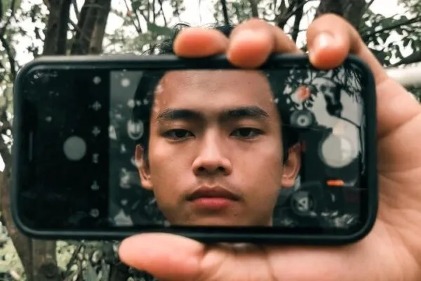 jovem olhando para a câmera através da tela do celular enquanto está na natureza, simbolizando equilíbrio e melhorar relação com a tecnologia