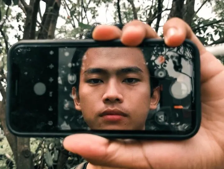 jovem olhando para a câmera através da tela do celular enquanto está na natureza, simbolizando equilíbrio e melhorar relação com a tecnologia