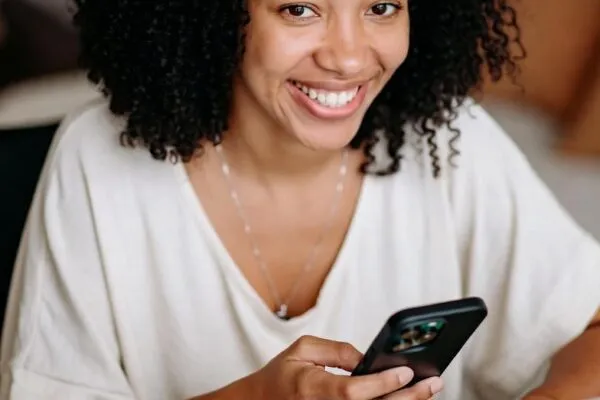Mulher brasileira sorrindo enquanto trabalha em casa com laptop e celular, com checklist, café e notas de real sobre a mesa.