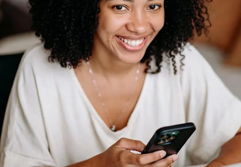 Mulher brasileira sorrindo enquanto trabalha em casa com laptop e celular, com checklist, café e notas de real sobre a mesa.
