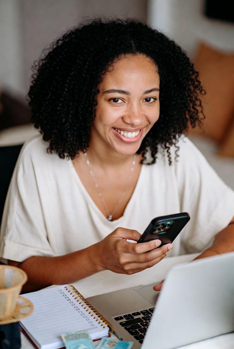 Mulher brasileira sorrindo enquanto trabalha em casa com laptop e celular, com checklist, café e notas de real sobre a mesa.